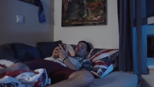 ベッドの上に横になっている間 彼のスマートフォンを見て退屈男 — ストック動画