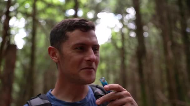 有魅力的年轻人一边在森林里从水合物背包里喝水 一边和他的徒步旅行伙伴一起笑着 — 图库视频影像