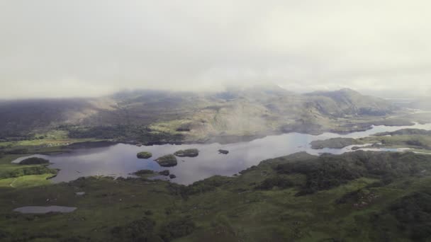 草で覆われた山々の間に位置する大きな湖と渓谷のアイルランドの美しい航空機 太陽の光が輝くムーディー雲の風景 雲の薄い層を後方に飛んで — ストック動画