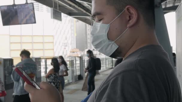 スカイトレインが接近するのを待っている携帯電話を使用している間 アジアの男は コロナウイルスCovid19からマスクを身に着けている — ストック動画