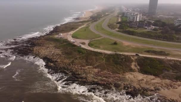 ウルグアイの霧の日の間にプンタ エステ市の海岸線に沿って海岸沿いの道路の壮大なドローンショット 崖に衝突する波 — ストック動画