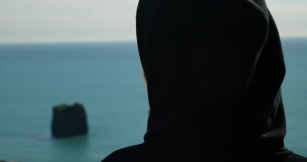 黒ずくめの長髪の少女がアイスランドの晴れた日に海を見て左と右を見ています — ストック動画