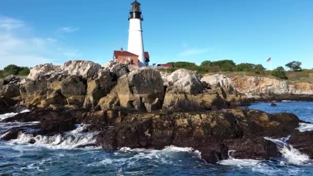 岩の多い海岸沿いのニューイングランド灯台 遠くにアメリカの国旗とのスローモーションで空中 旅行や観光のテーマ ポートランド メイン — ストック動画