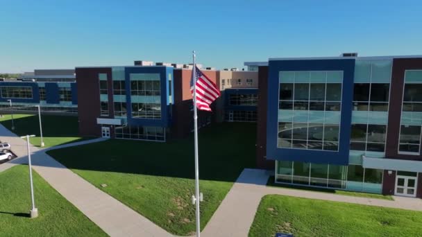 空中ドローン映像はアメリカの公立学校の前で手を振るアメリカの旗を特徴としています 近代的で新しい校舎には大きな窓と歩道がある アメリカの教育のテーマ — ストック動画