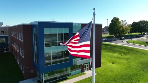 在学校大楼前 旋转的美国国旗在风中飘扬 阳光明媚的早上 当地的公立高中有很大的玻璃窗和新教室 — 图库视频影像