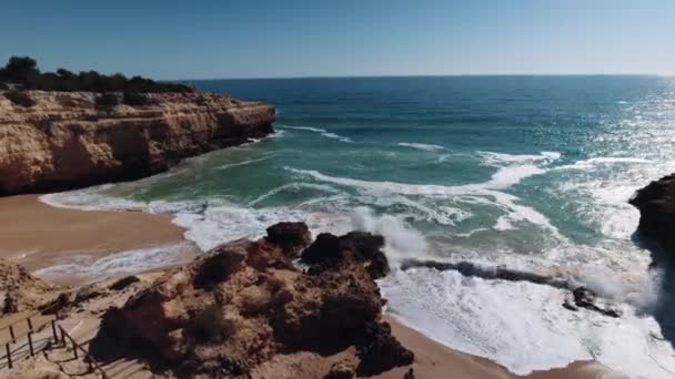 ポルトガルのアルガルヴェのアルバ リゾートで岩に衝突する波 背景に美しい海と空 海の向こうのビーチから飛んでくるドローン映像 カメラの下の水が画面を埋め — ストック動画