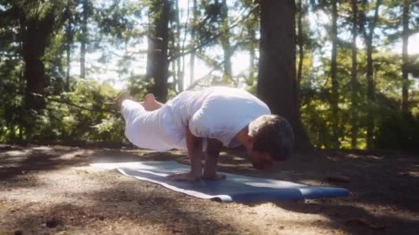 近距离拍摄的高加索成年男子身穿白色衣服 持有高级瑜伽姿势 孔雀蛋黄酱 在松树林 相机运动 — 图库视频影像