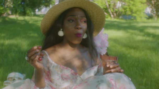 公園でピクニックブランケットで笑顔を楽しむ黒の女性食べるブドウ — ストック動画