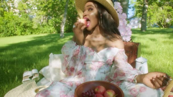 Smiling Black Woman Park Picnic Floral Dress Takes Bite Apple — Vídeo de stock