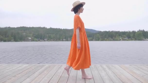 美しい女の子で活気のあるオレンジのドレス湖畔の木製デッキを歩く — ストック動画