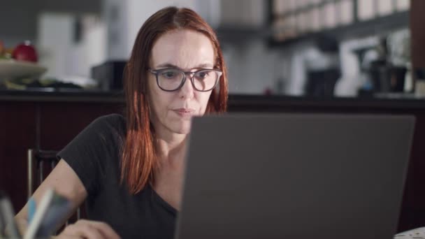 赤髪の熟女は彼女の台所の横に家に座っています 読書と彼女のラップトップコンピュータ上で閲覧 — ストック動画