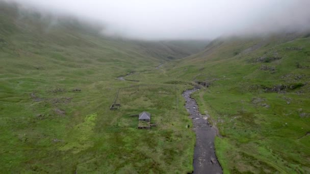 空中无人驾驶飞机镜头缓慢的向后飞行 并通过苏格兰冰川中的大气低垂的云彩下降 以揭示一条河流和Gameshope Bothy 一座山区避难所 苏格兰 — 图库视频影像