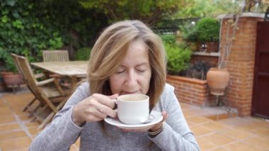Olgun beyaz kadın bahçesinde rahatça bir fincan kahve içiyor.