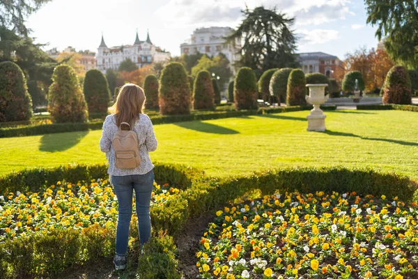 Halk parkında güzel çiçek tarhları olan bir kadın, yemyeşil bitki örtüsünü düşünüyor.