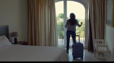 Elinde bavuluyla otel odasına gelen bir kadın balkon manzarasına hayran olmak için kapıları açıyor.