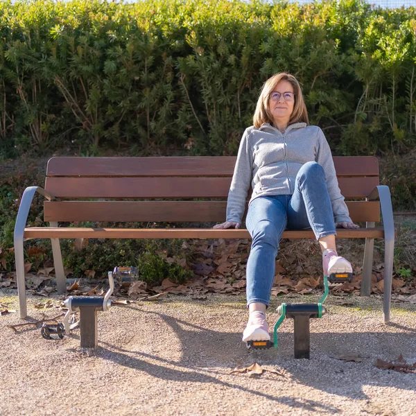 Mature woman sitting on a bench and pedaling on a gymnastics apparatus in a public park
