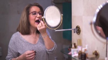 Older white woman painting her lips in front of the mirror of her bath