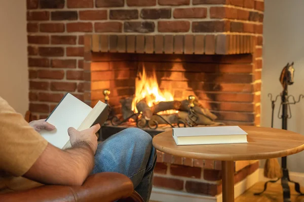 Man sitting in an armchair and reading a book in a relaxed way in front of the fireplace fire in winter