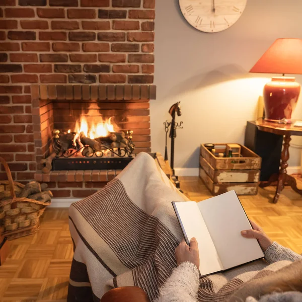 Woman sitting in a chair, covered with a blanket and reading a book comfortably in front of the fireplace fire
