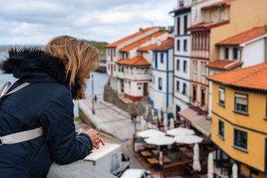Sokak balkonundan sarkan ve Cudillero, Asturias 'taki balıkçı köyünün manzarasını seyreden turist kadın.
