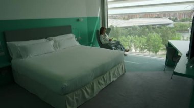 Koltukta oturan kadın, odanın geniş bir penceresinden şehrin manzarasını seyrediyor.