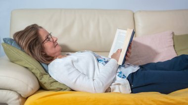 Kanepede uzanmış, mutlu ve rahat bir tavırla kitap okuyan beyaz kadın..