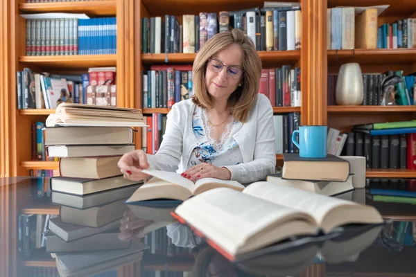 stock image Senior white woman reading a book on a glass table with a bookcase full of book behind her