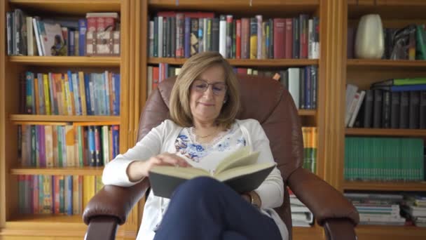 年长的白人妇女坐在一张放松的扶手椅上看书 书架在她身后 — 图库视频影像