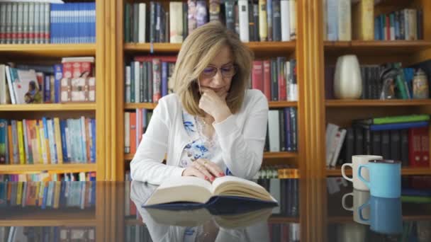 Weiße Seniorin Sitzt Vor Einem Glastisch Und Liest Ein Buch — Stockvideo