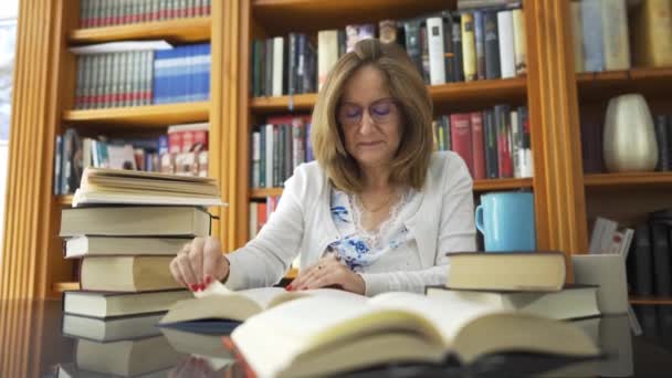 一个白人妇女坐在玻璃桌旁工作 身边和书架上都有很多书 — 图库视频影像