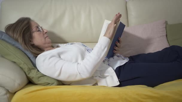 一位年长的白人妇女躺在沙发上 以一种放松和休息的态度看书 — 图库视频影像