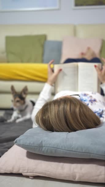 女人一边翻书 一边和她的狗躺在地板上的毛毯上 — 图库视频影像