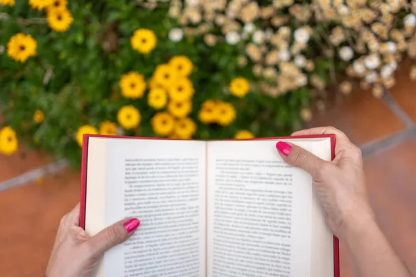 Womans hands on a book while reading in the garden next to flowering plants