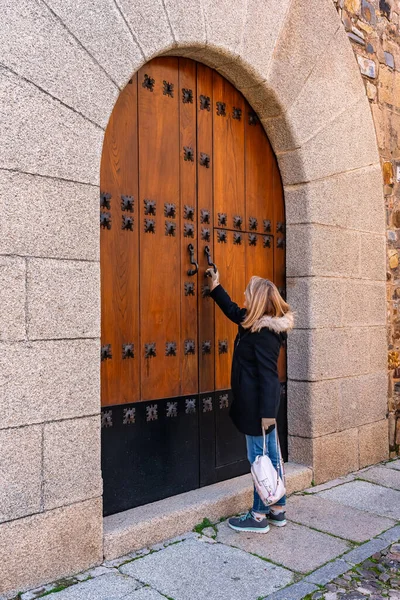 Woman knocking on a large door in a medieval building in the city of Caceres
