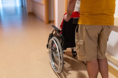 Arkadan gelen bir adam, tekerlekli sandalyede oturan yaşlı bir insanı iç koridorda gezdiriyor.