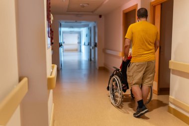 Yaşlı bir adam ve kadının tekerlekli sandalyelerinde yürüdüğü uzun bir koridor.