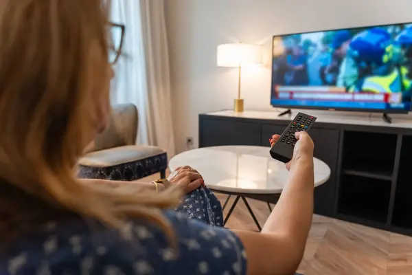 Sırtı ona dönük bir şekilde koltukta oturan ve uzaktan kumandayla televizyon kanalını değiştiren bir kadın..