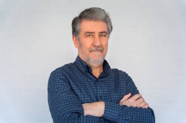 Beyaz, yaşlı, sakallı ve kollarını kavuşturmuş bir adam kameraya bakıyor..