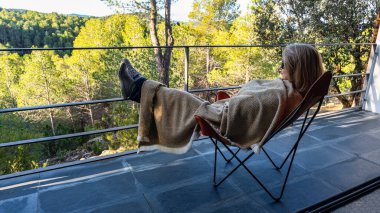 Sandalyeye uzanmış, battaniyesini sarmış, terastan orman manzarasının keyfini çıkaran bir kadın.