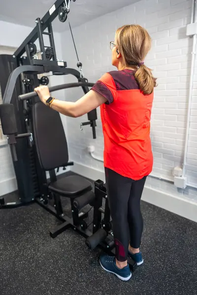 Olgun bir kadın evinde ağırlık makinesiyle güç egzersizleri yapıyor.