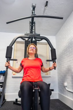 Vücut geliştirme egzersizi yapan olgun bir kadın evinde ağırlık makinesiyle göğüs kafesi yapıyor.