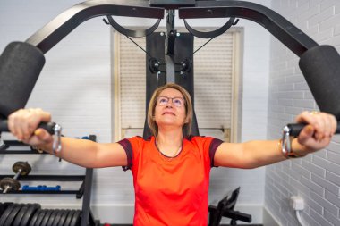 Vücut geliştirme egzersizi yapan olgun bir kadın evinde ağırlık makinesiyle göğüs kafesi yapıyor.