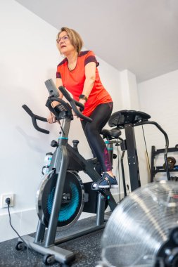 Evde spor salonunda egzersiz yapan yaşlı bir kadın menopozun sıcaklığı için vantilatörü açık..