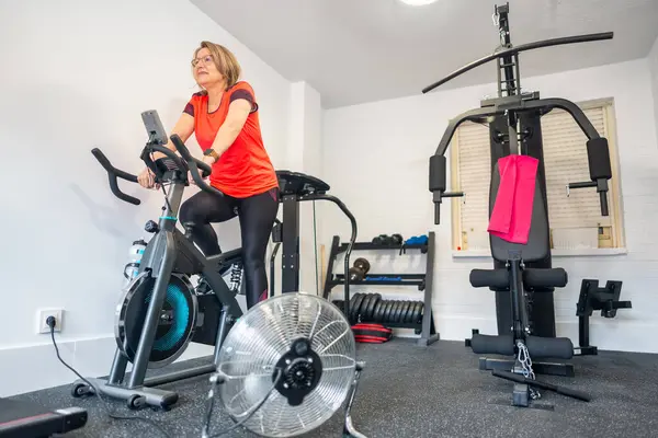 Evde spor salonunda egzersiz yapan yaşlı bir kadın menopozun sıcaklığı için vantilatörü açık..