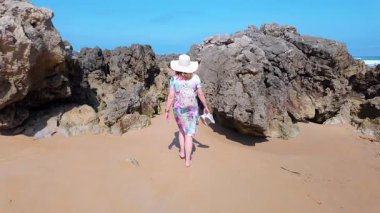 İspanya 'nın kuzeyinde, Kosta Quebrada, Cantabria' da, yalnız bir kumsalda gezen elbiseli ve şapkalı turist kadın.