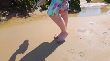 Sahilde kumsalda yürüyen ve ayaklarını küçük su havuzlarına koyan çıplak ayaklı bir kadın..
