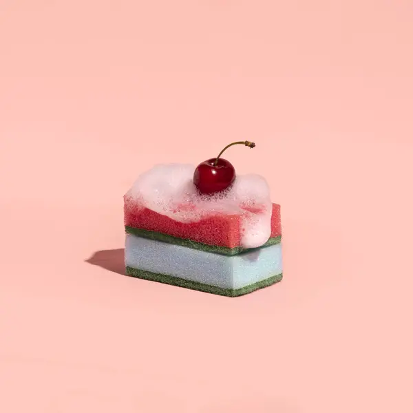 Dishwashing sponges, stacked like a creamy cake, foam over them and a cherry on top. Creative dessert layout, dusty pink background.