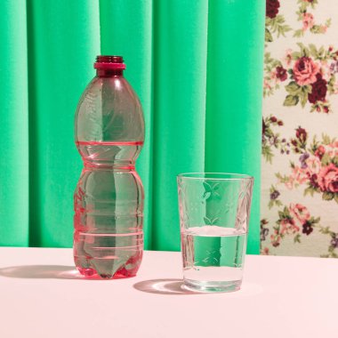 Plastik şişe, bir bardak içme suyu, retro tarzı çiçekli duvar kağıdı ve zümrüt yeşili kumaş..