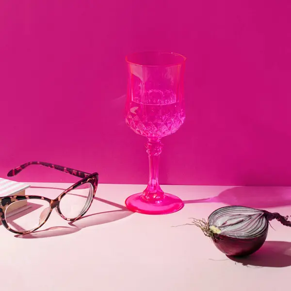 Cocktail drink, reading glasses and fresh purple onion, creative aesthetic still life. Fuchsia pink wall in background.