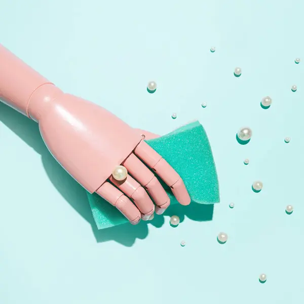 Wooden pink painted hand holding a sponge, pearl beads spilled against pastel blue green background. Creative cleaning concept.